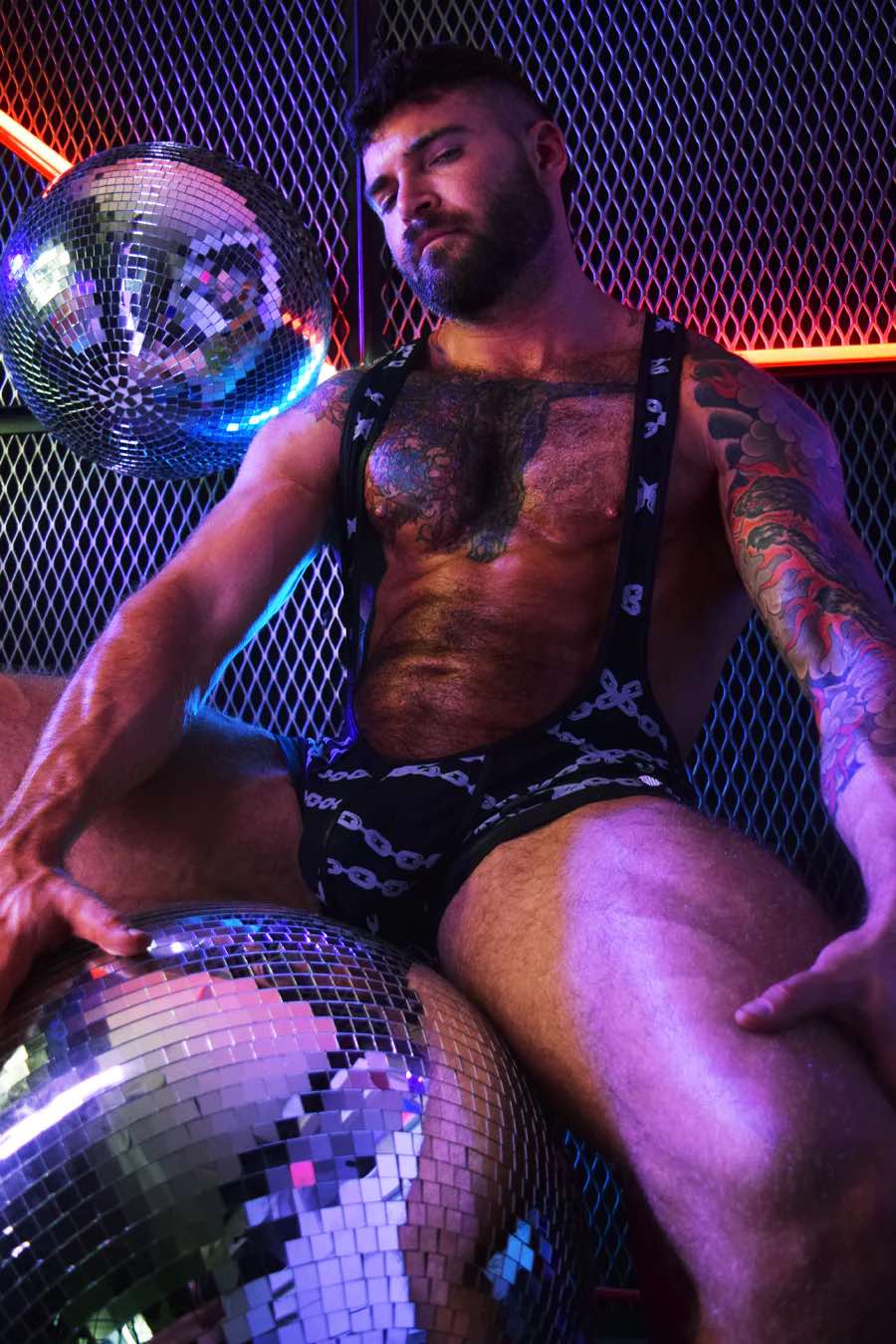A model leaning next to a neon fence and sitting on a disco ball wearing the Dirty Boy Chains Singlet.