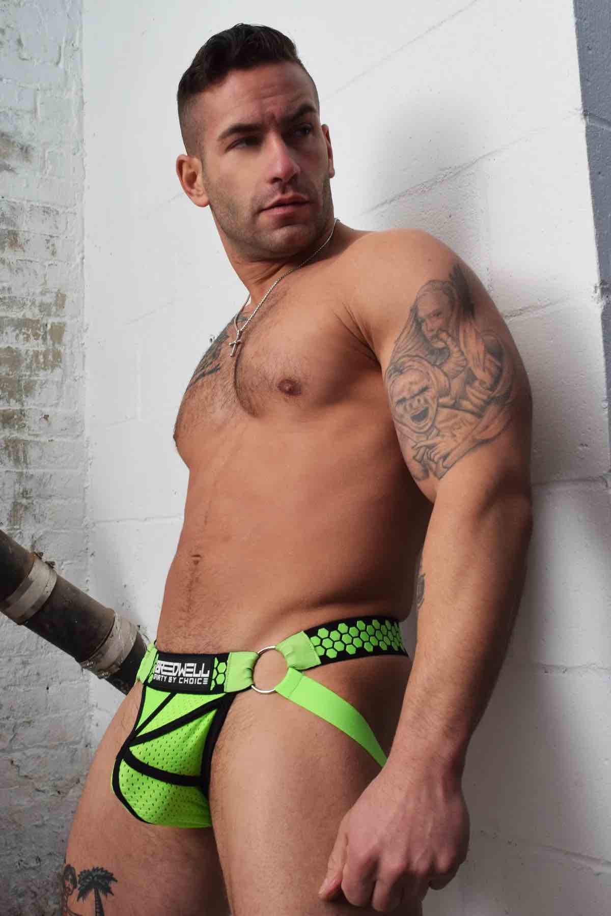 A model leaning against a white brick wall wears the neon green Hex Jock, front view.