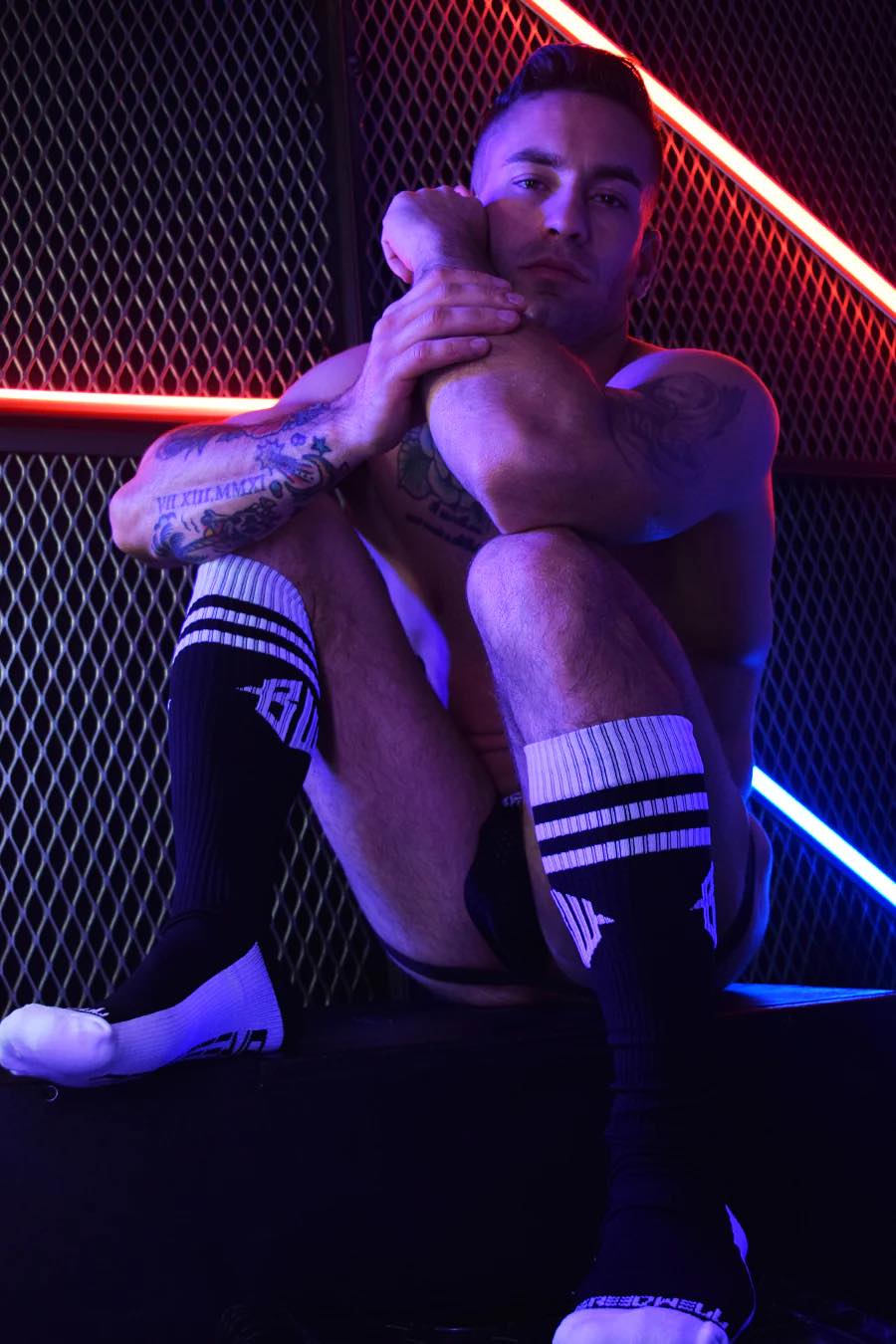 A model in a jock strap sitting in front of a fence with neon lights wearing the black Hex Socks.