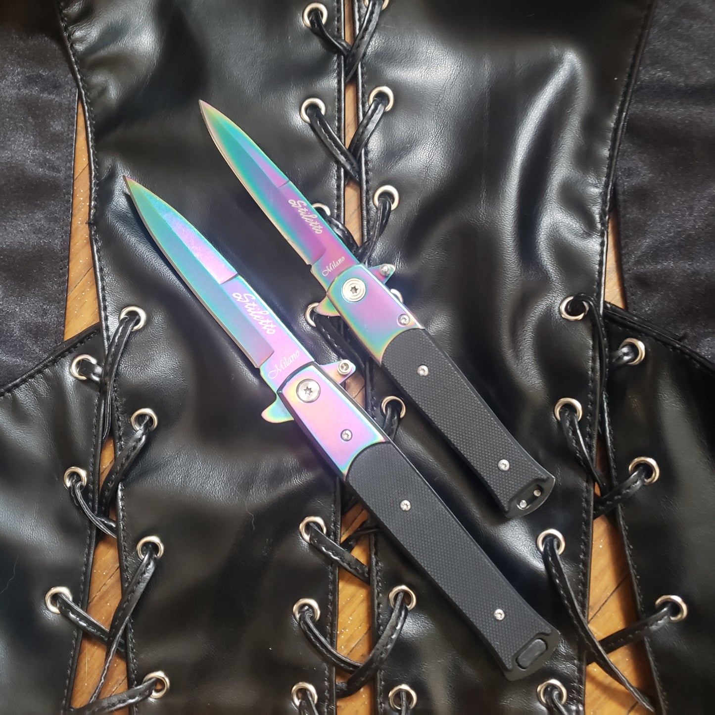 One 4" and one 5" rainbow with black handle Stiletto Type Folding Knife in the open position laying on a leather vest.