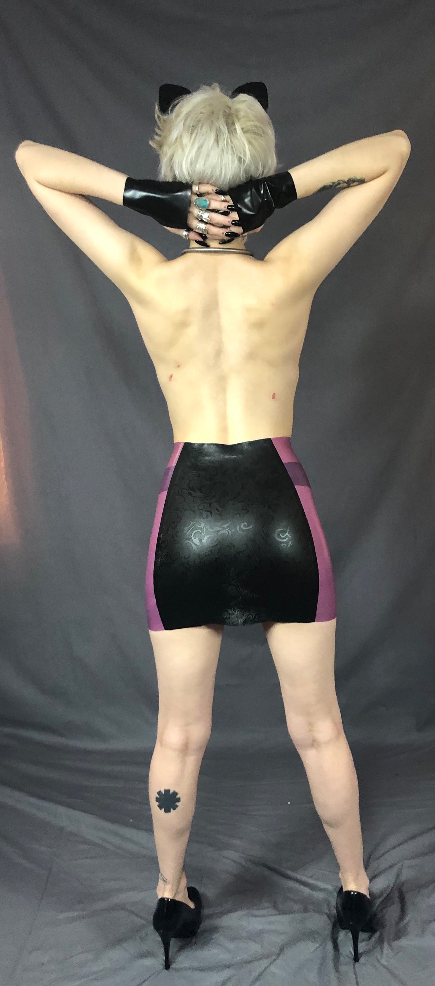A feminine model wearing cat ears, holding black latex gloved hands across the back of their neck and showing the back of the black and magenta Latex Girdle Mini Skirt.