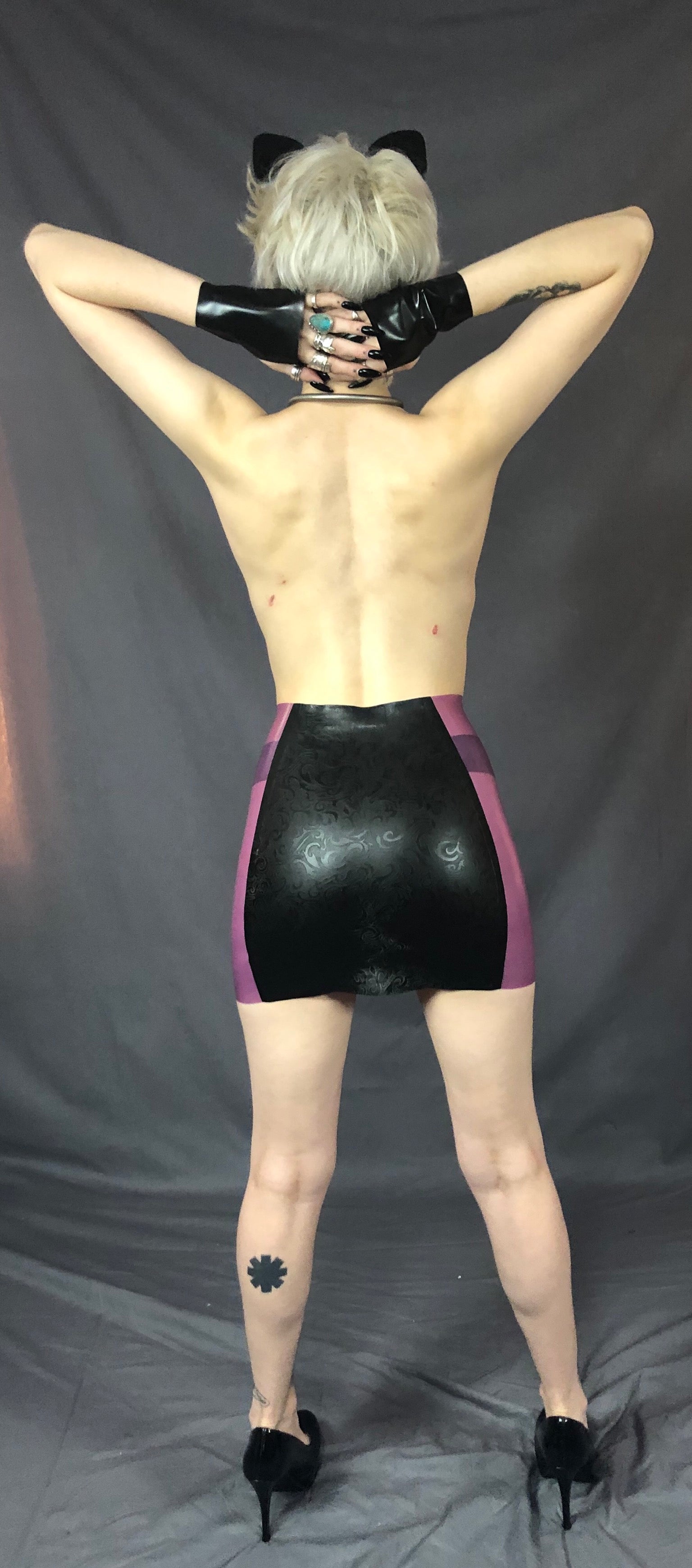 A feminine model wearing cat ears, holding black latex gloved hands across the back of their neck and showing the back of the black and magenta Latex Girdle Mini Skirt.