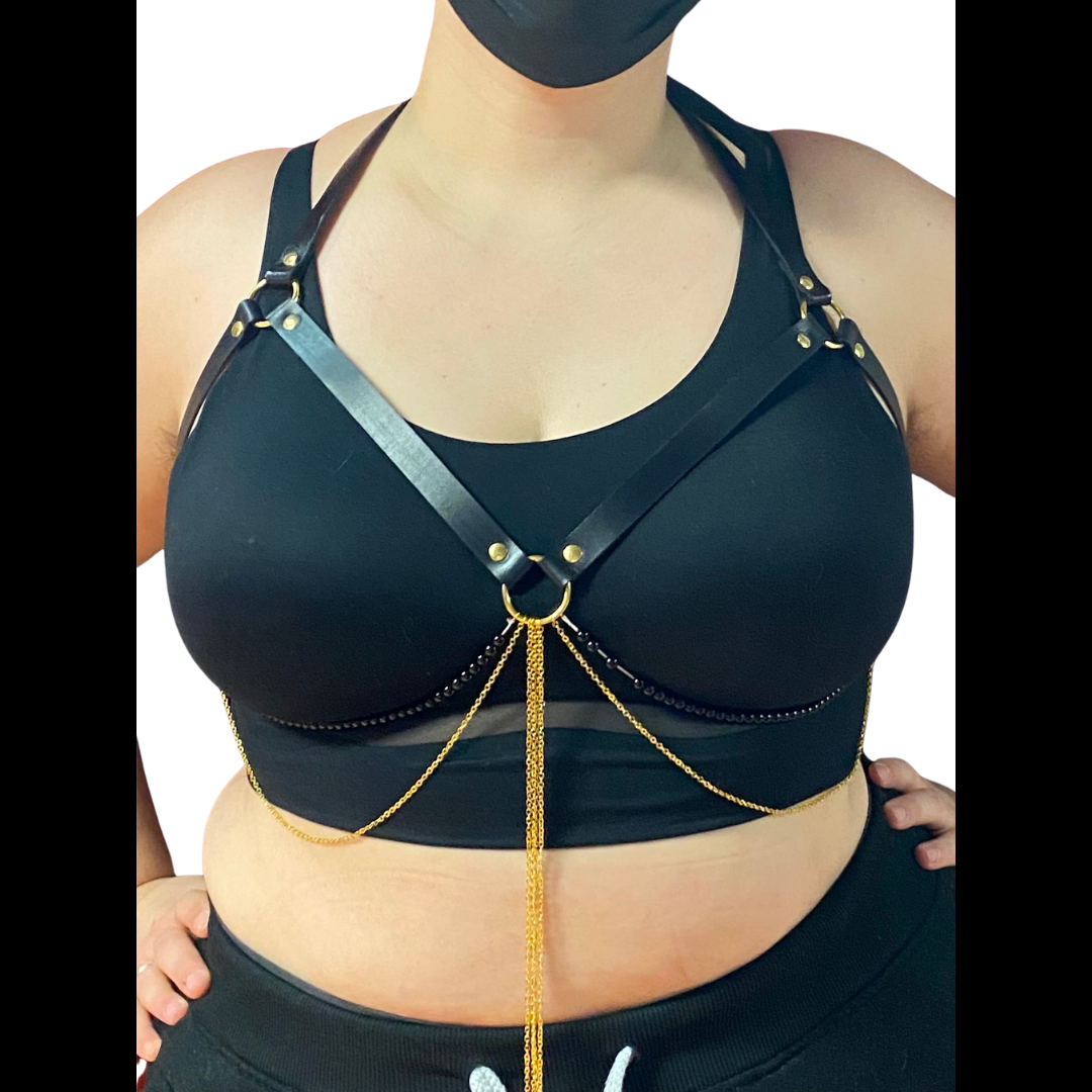 A curvy model in a black bra models the Jewel Harness in Onyx from the front. The leather straps form a halter that frames their breasts, with two strings of black beads and several fine gold-colored chains hanging from the center ring and tracing the underside of their breasts. It fastens with gold-colored hardware.