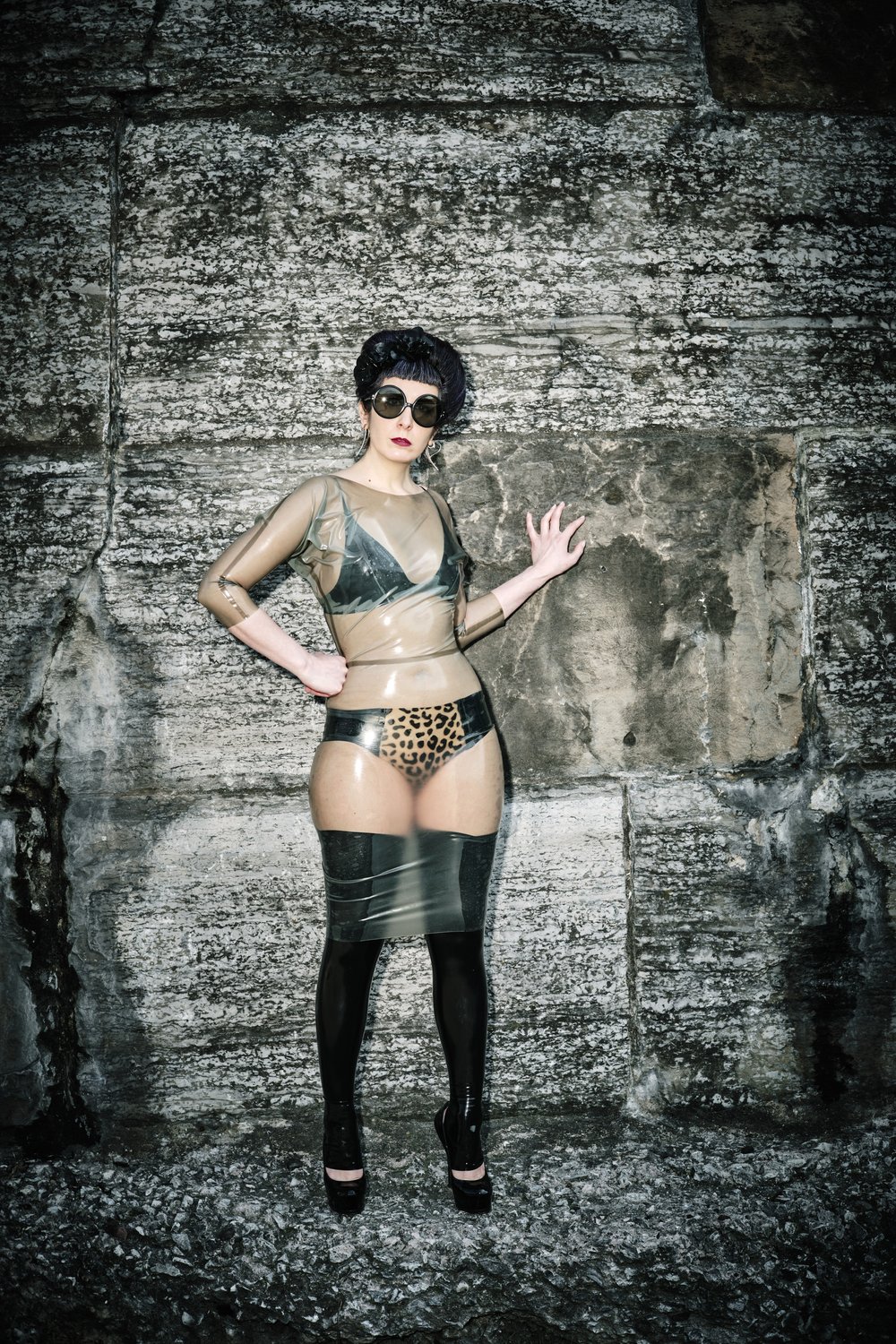 A model standing against a stone wall wearing the black and leopard print Specialty Latex Panel Panty with a latex bra under a sheer latex dress.