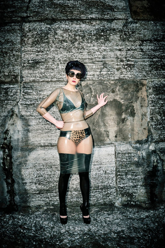 A model standing against a stone wall wearing the black and leopard print Specialty Latex Panel Panty with a latex bra under a sheer latex dress.