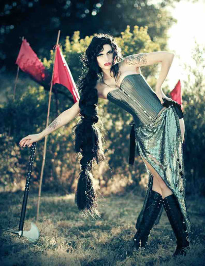 A model at a ren faire posing with an axe wears the Gray Taffeta Slim Corset over a shimmery skirt.