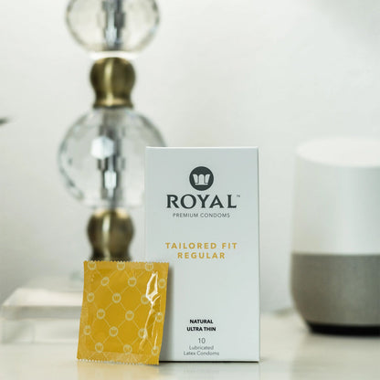 A 10 pack box of Tailored Fit Regular Royal Condoms with a single condom in a yellow packet leaning up against it and a lamp in the background.