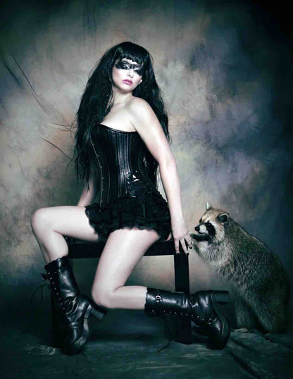 A model sitting on a bench next to a racoon, wearing the Black Leather Short Overbust Corset -Slim over ruffled shorts.