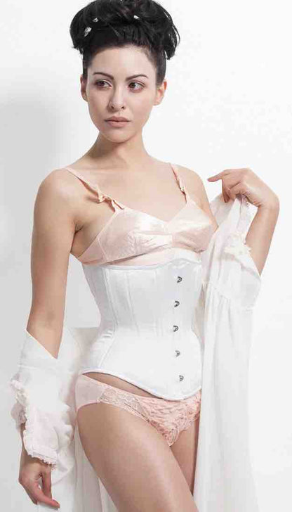 A model wearing the White Satin Mid-Length Underbust Corset - Hourglass over a pastel pink bra and panties.