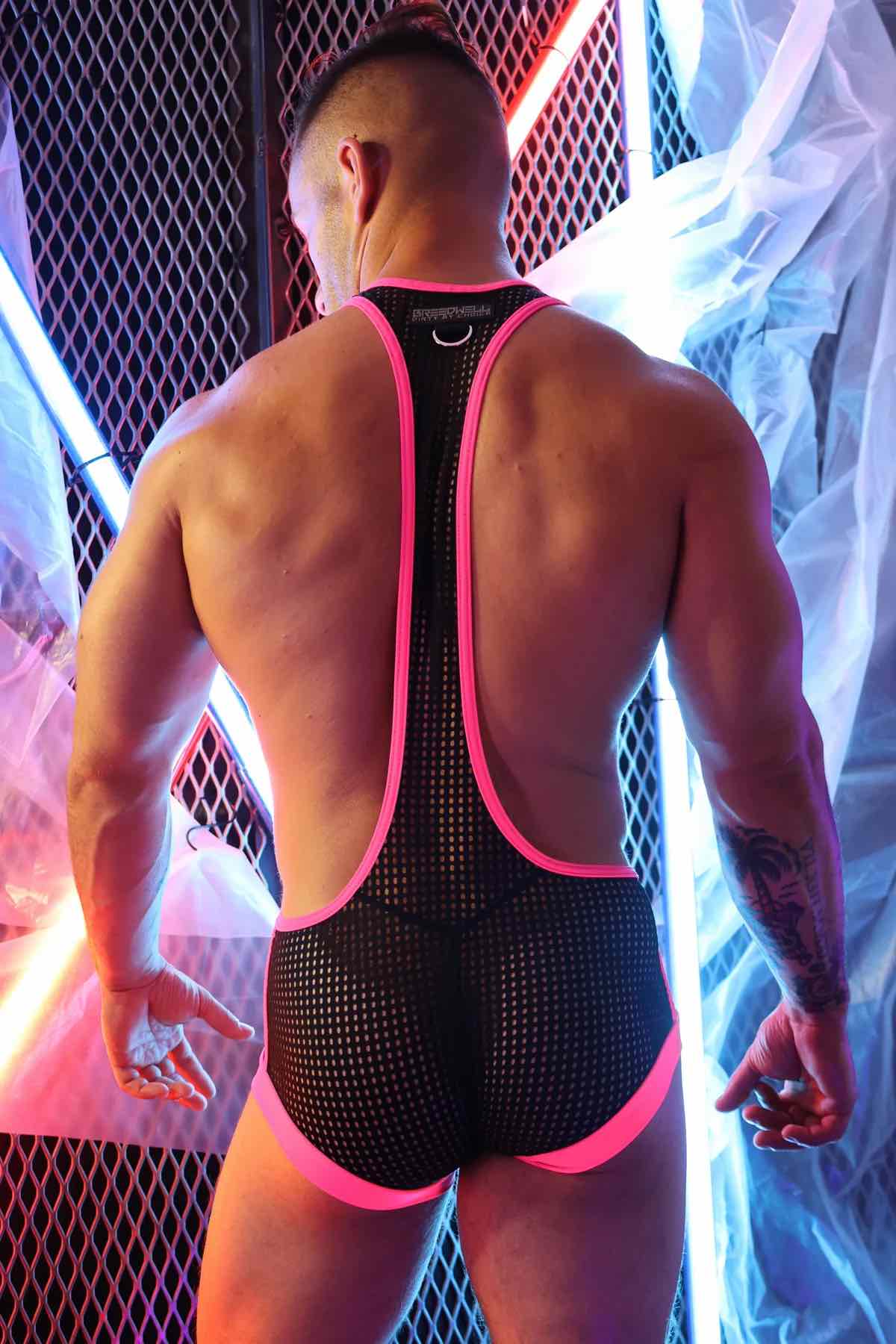 A model wearing the pink and black Dirty Boy Neon Trim Singlet against a fence lit with neon lights, rear view.