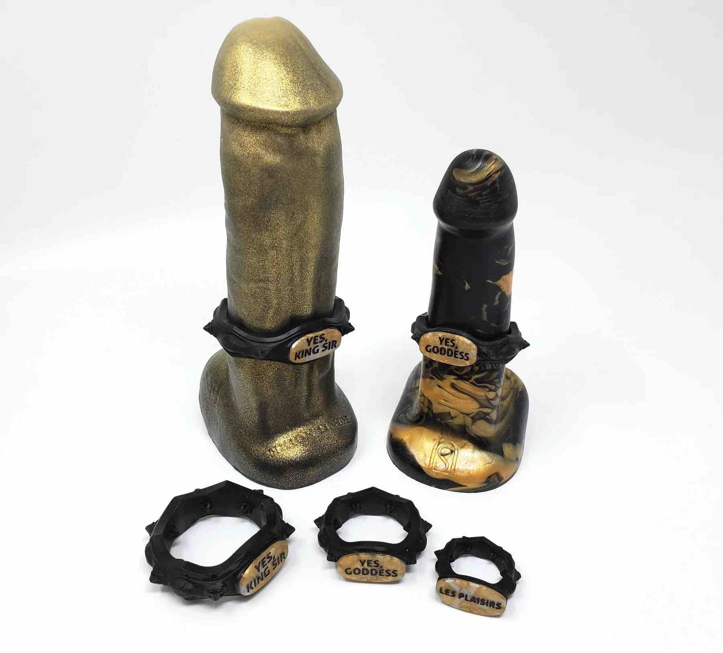 Three black and gold Royal Fetish FEMDOM Edging Body Bands sitting in front of two black and gold dildos with body bands on them.