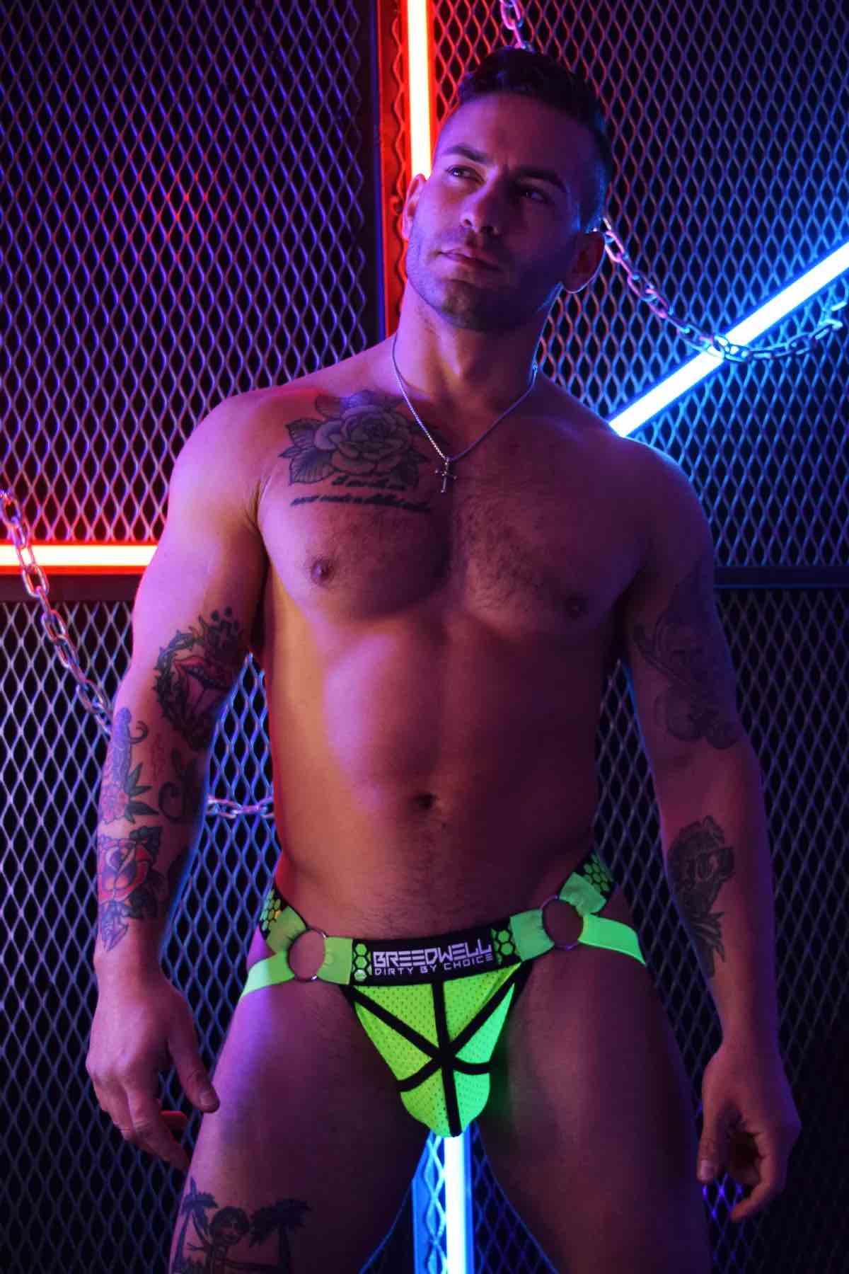 A model in front of a fence with neon lighting wears the neon green Hex Jock, front view.