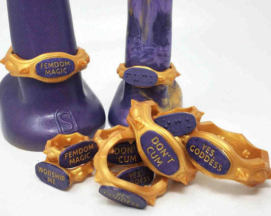 A pile of gold and purple Royal Fetish FEMDOM Edging Body Bands in front of two dildos donned with body bands. They show different messages in text and braille. 