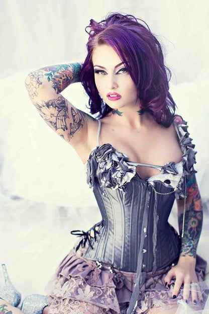 A tattooed model with purple hair wears the Gray Taffeta Slim Corset over a grey ruffled bra and purple ruffled skirt,