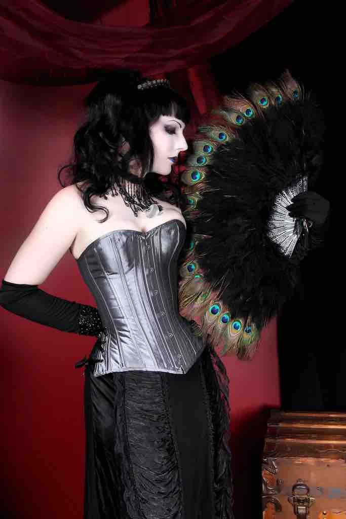 A model standing in front of a red curtain wearing black gloves and holding a peacock feather fan wears the Gray Taffeta Slim Corset over a black skirt.