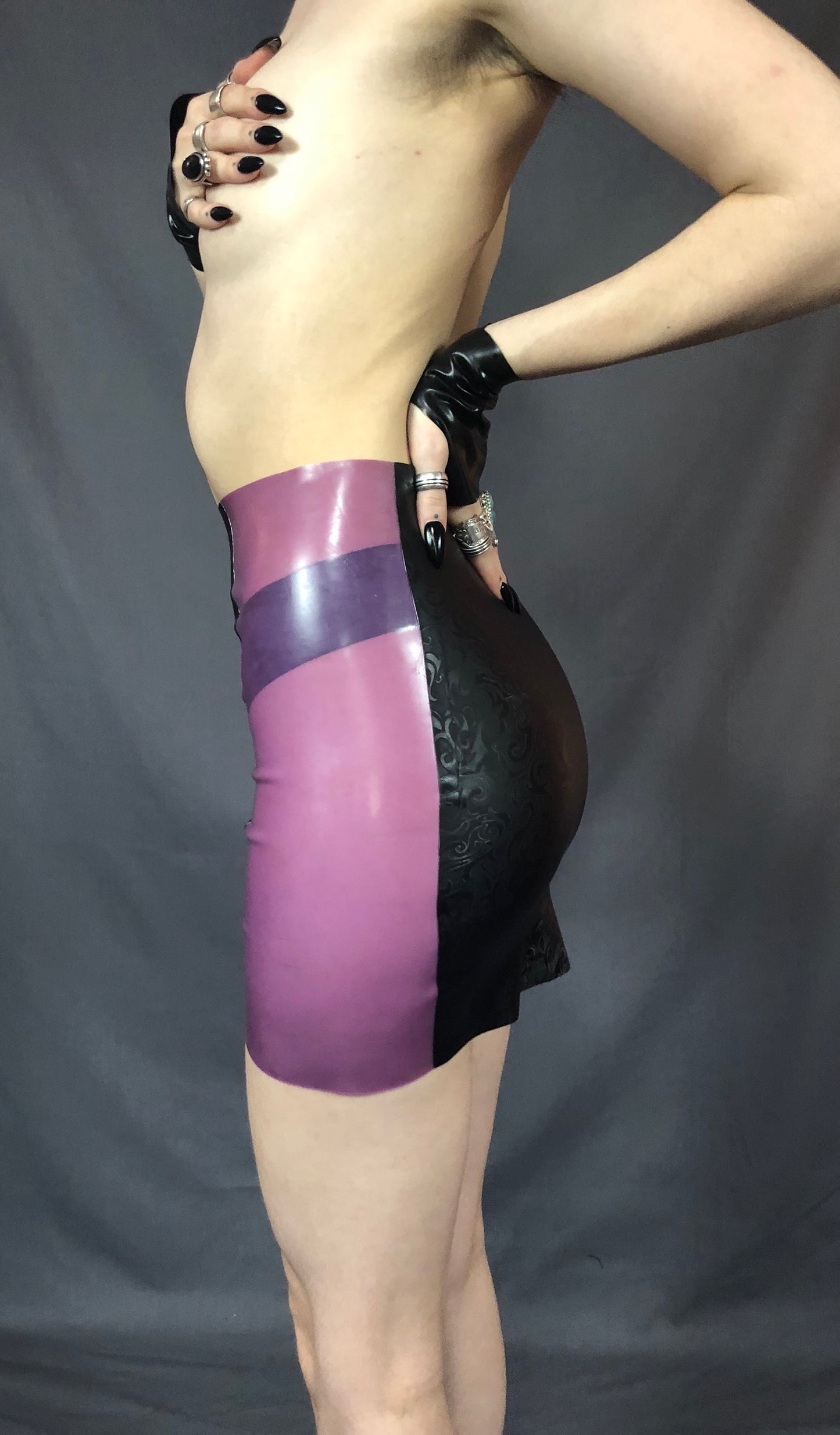 A feminine model holding a black latex gloved hand over their breasts showing the left of the black and magenta Latex Girdle Mini Skirt.