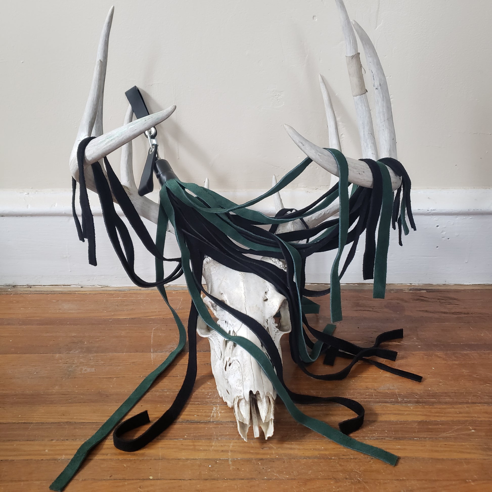 The green and black suede finger loop flogger displayed on the antlers of a deer skull.