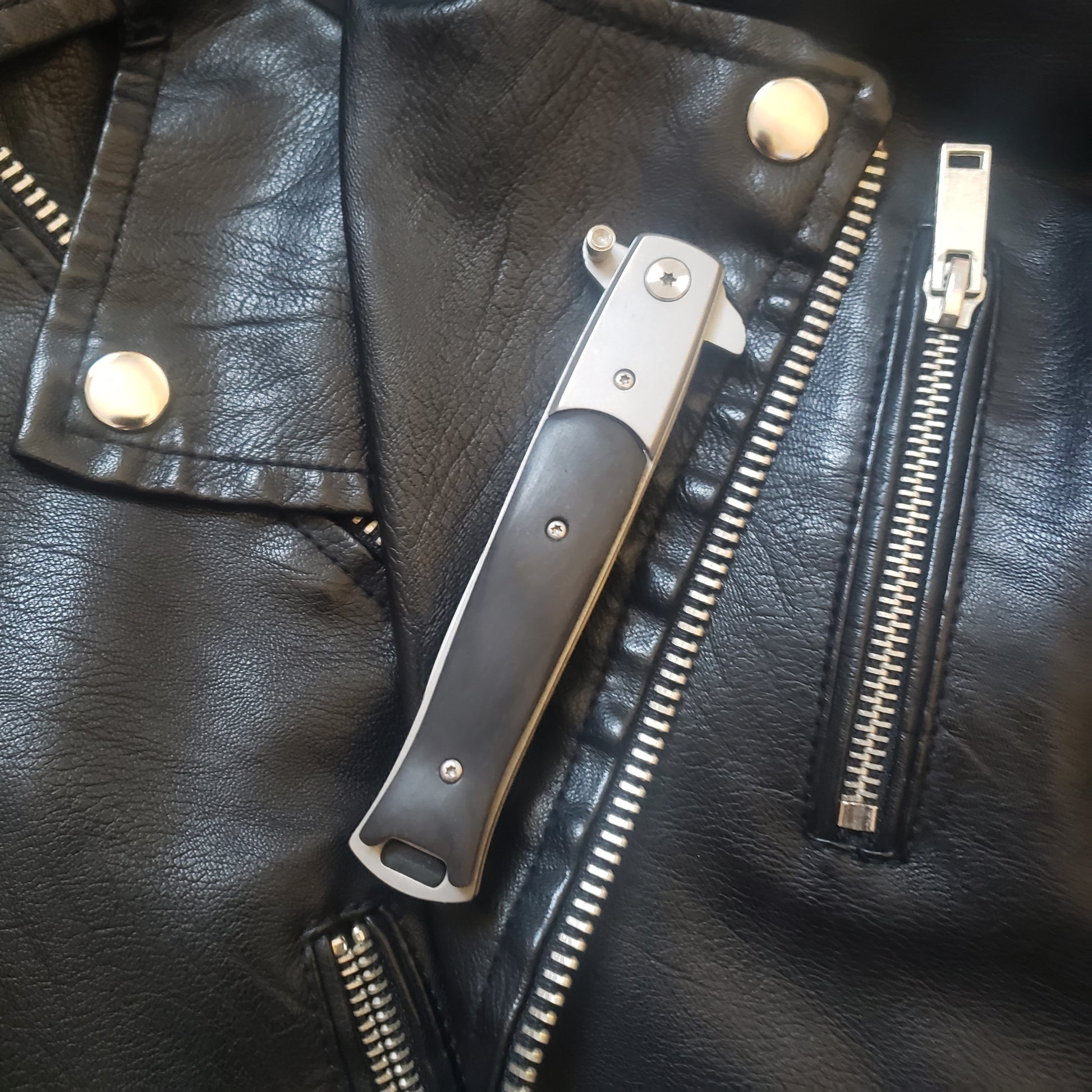 A folded black Stiletto Type Folding Knife laying on a leather jacket. 