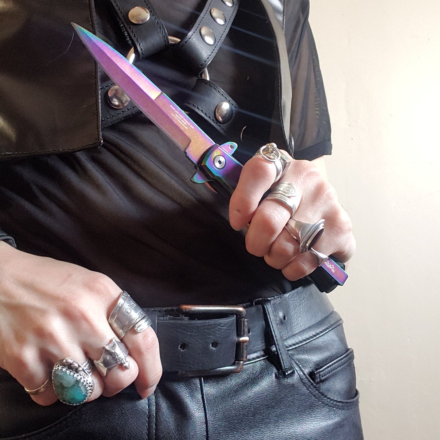 A model in a leather vest, black t-shirt and harness holding the black and rainbow Stiletto Type Folding Knife.