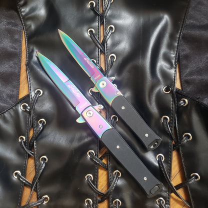 One 4" and one 5" rainbow with black handle Stiletto Type Folding Knife in the open position laying on a leather vest.