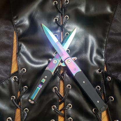 One 4" and one 5" rainbow with black handle Stiletto Type Folding Knife in the open position with the blades crossed against each other laying on a leather vest.