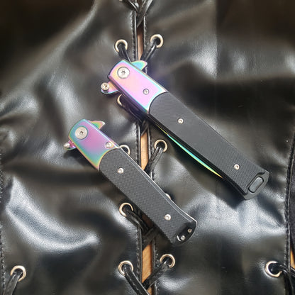 One 4" and one 5" rainbow with black handle Stiletto Type Folding Knife in the closed position laying on a leather vest.