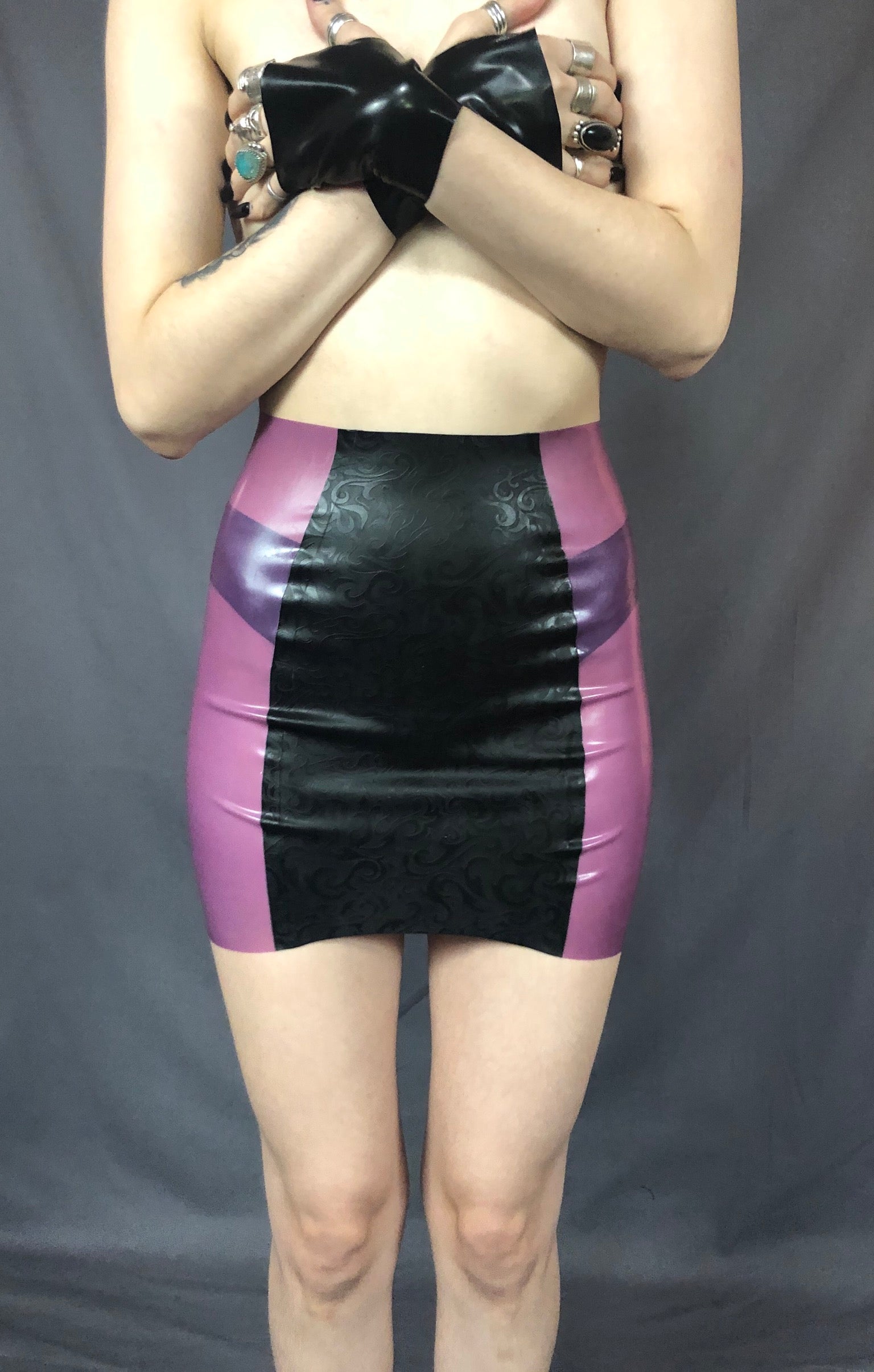 A feminine model holding black latex gloved hands over their breasts showing the front of the black and magenta Latex Girdle Mini Skirt.