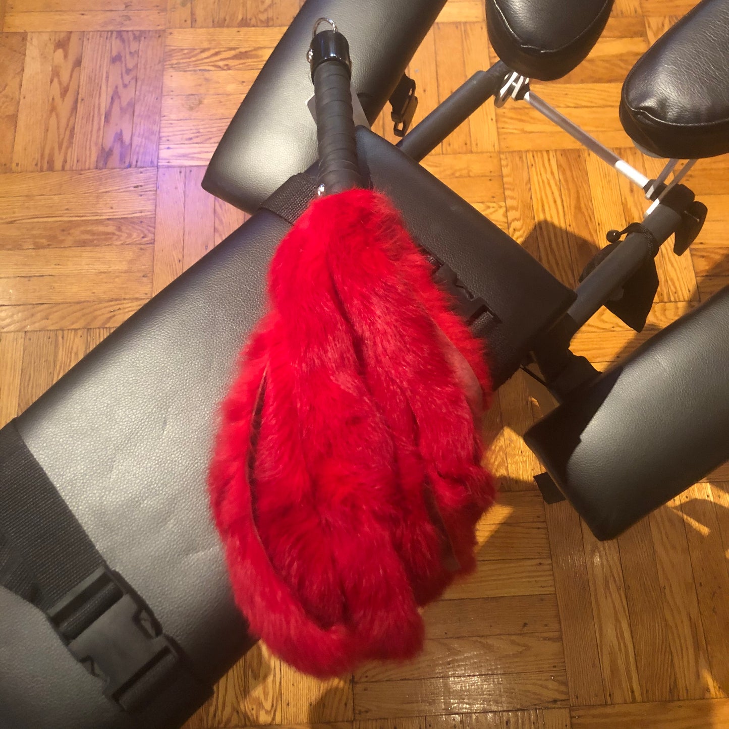 Red 20" rabbit fur flogger with black leather handle and D-ring for hanging, laying on a spanking bench.