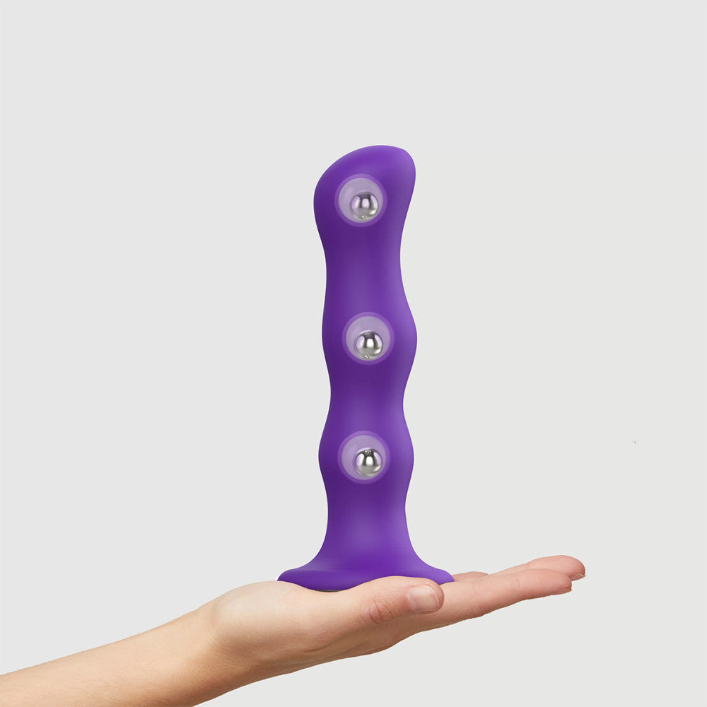 A model's hand holding up the purple Strap-On-Me Shaking Balls Dildo. An illustration shows the placement of the balls inside the dildo.