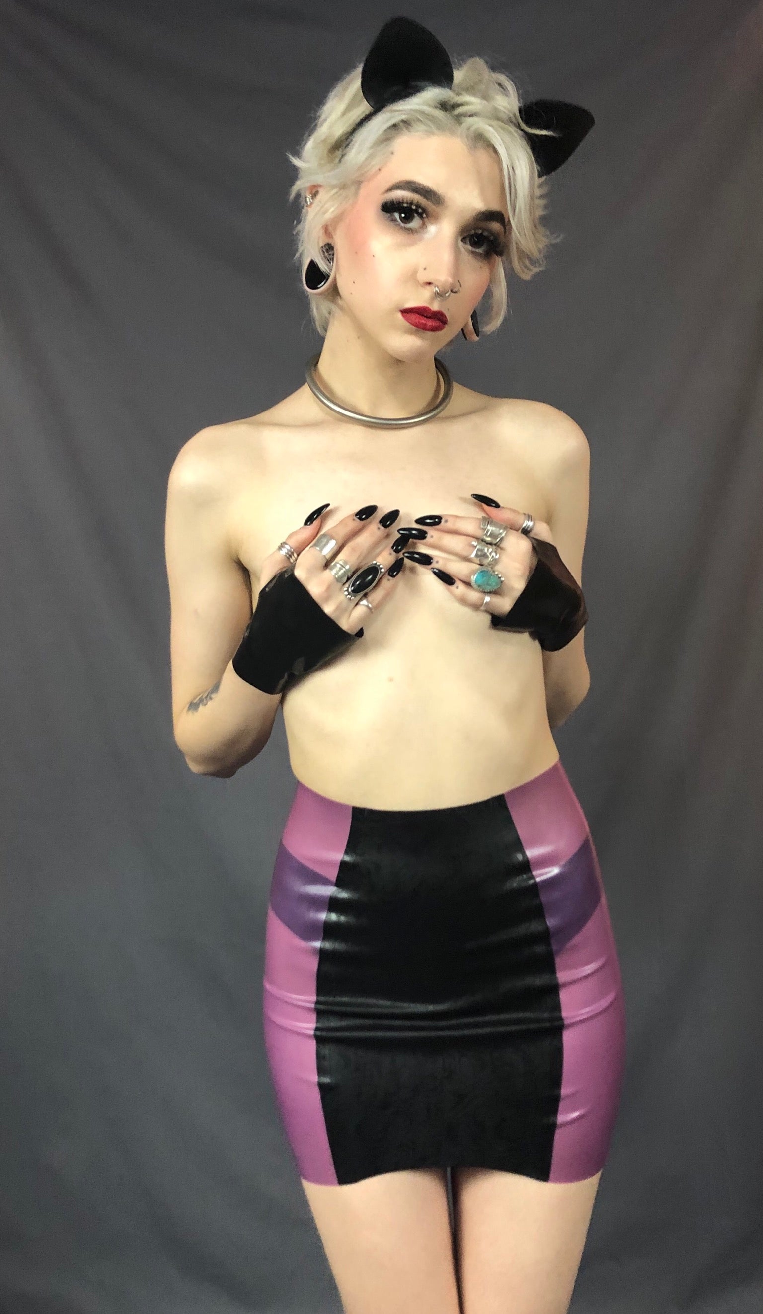 A feminine model wearing cat ears, holding black latex gloved hands over their breasts and showing the front of the black and magenta Latex Girdle Mini Skirt.