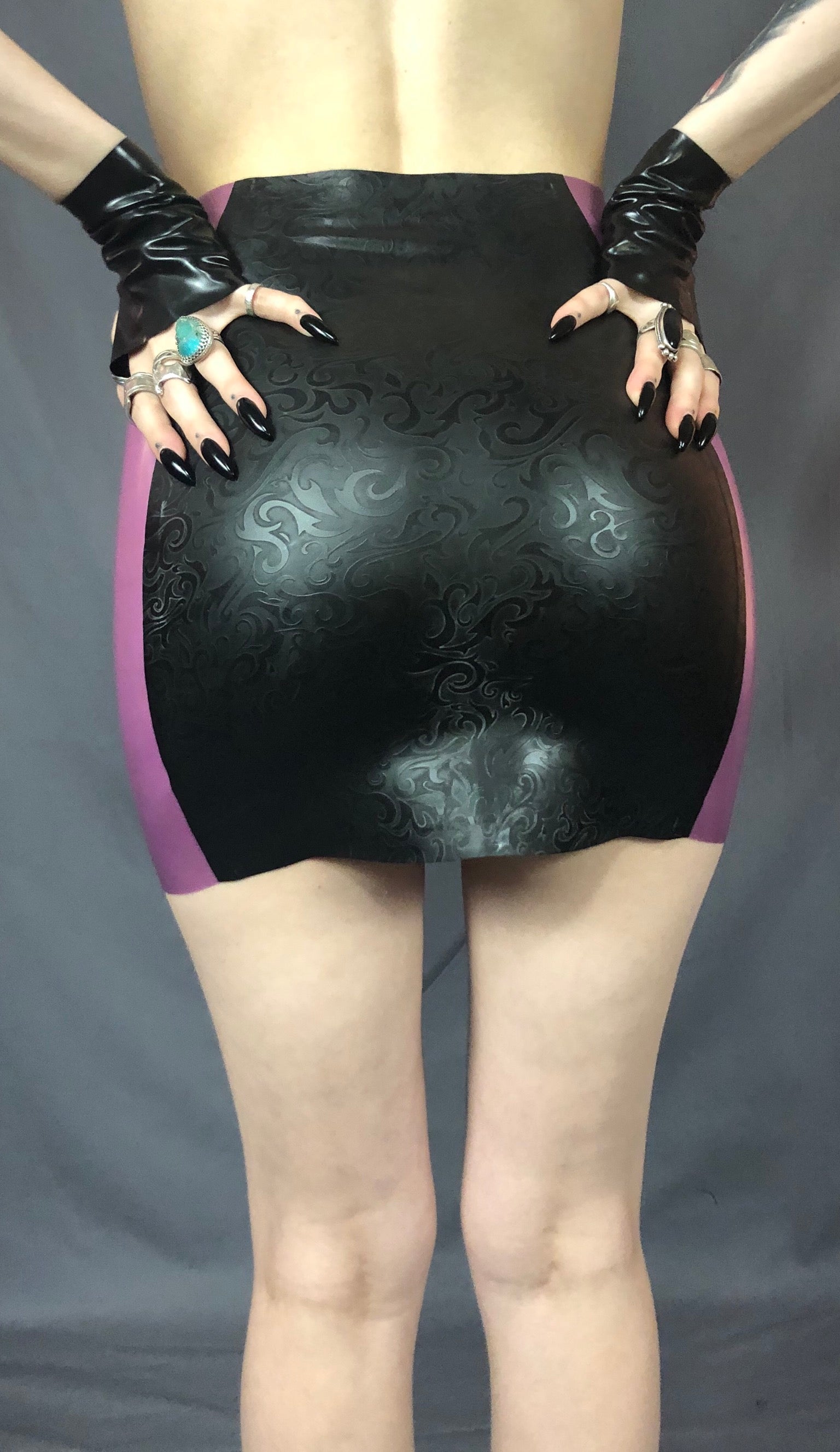 The lower torso of a model with latex gloved hands on their hips shows the back of the Latex Girdle Mini Skirt.