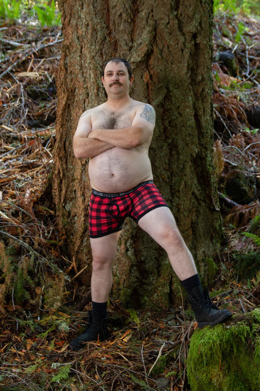 A masculine looking model standing shirtless in front of a tree in the forest wears the Backwoods Boxer Briefs. 