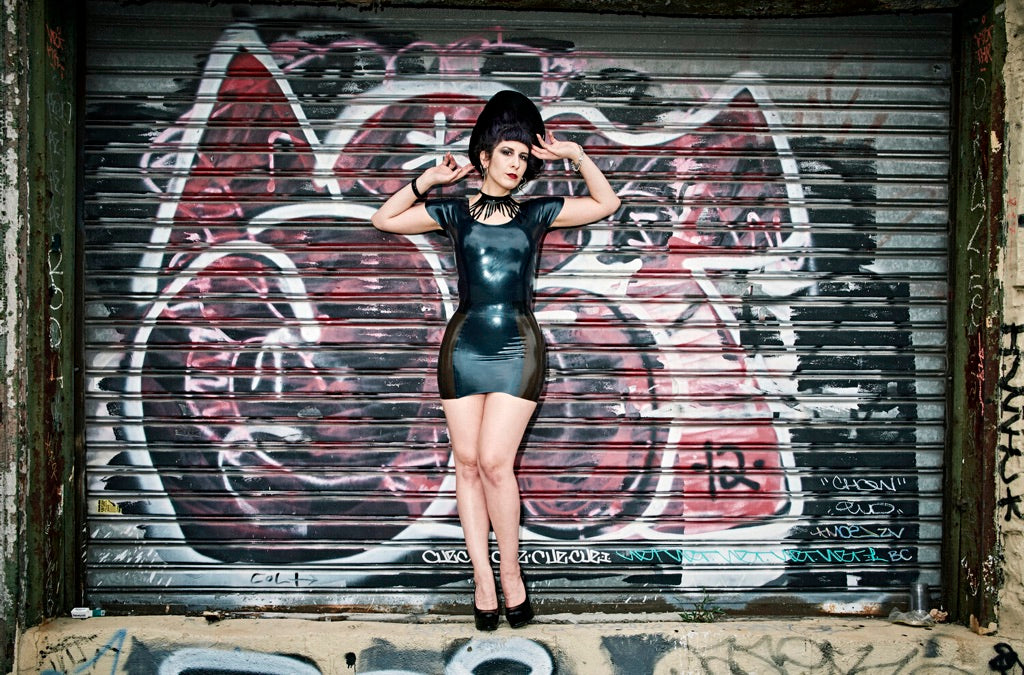 A model standing in front of a graffitied garage door showing the front of the black and black transparent Latex Girdle Mini Skirt with matching short sleeved latex top.