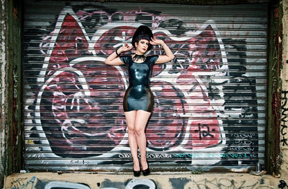 A model standing in front of a graffitied garage door showing the front of the black and black transparent Latex Girdle Mini Skirt with matching short sleeved latex top.
