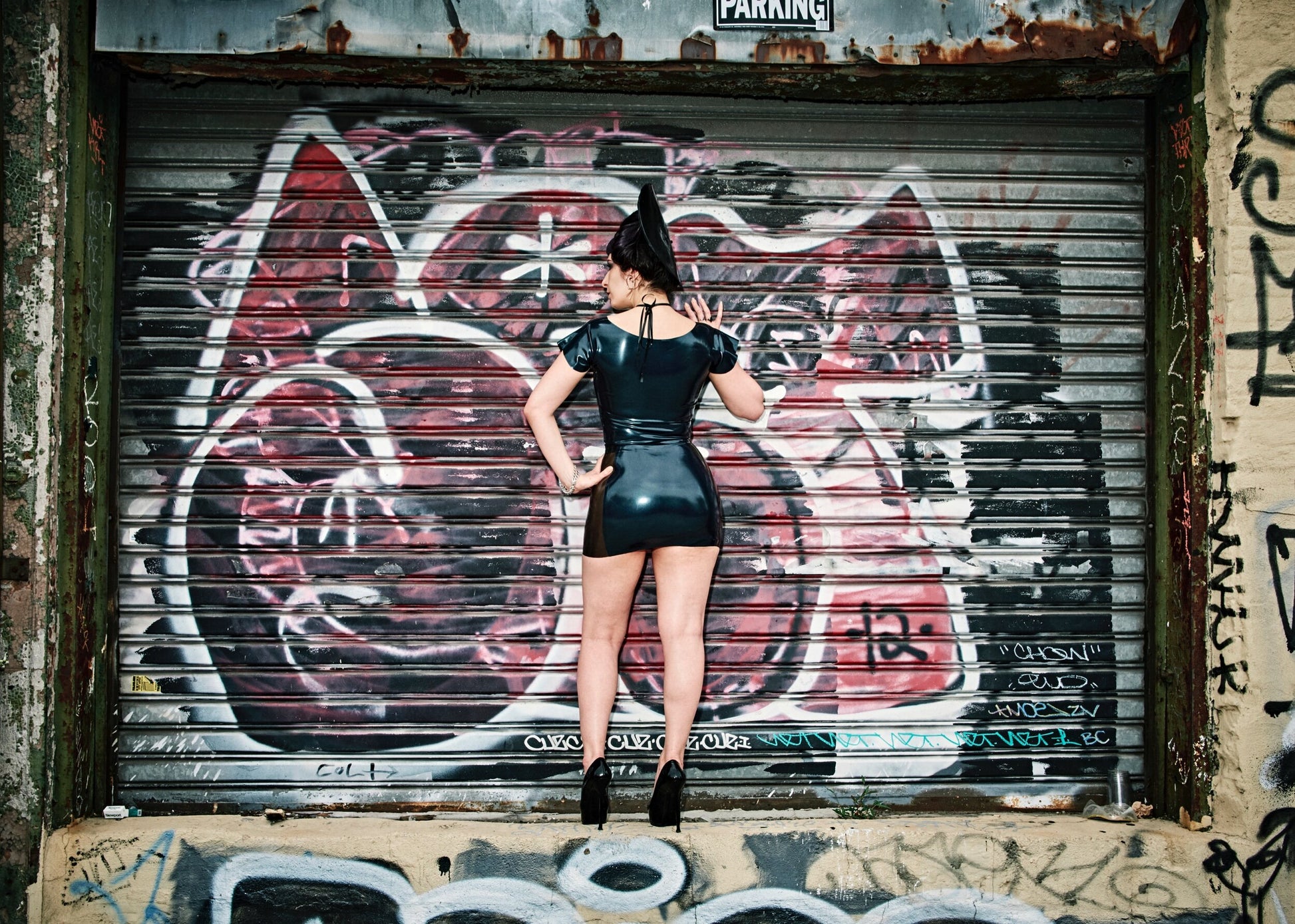 A model standing in front of a graffitied garage door showing the back of the black and black transparent Latex Girdle Mini Skirt with matching short sleeved latex top.
