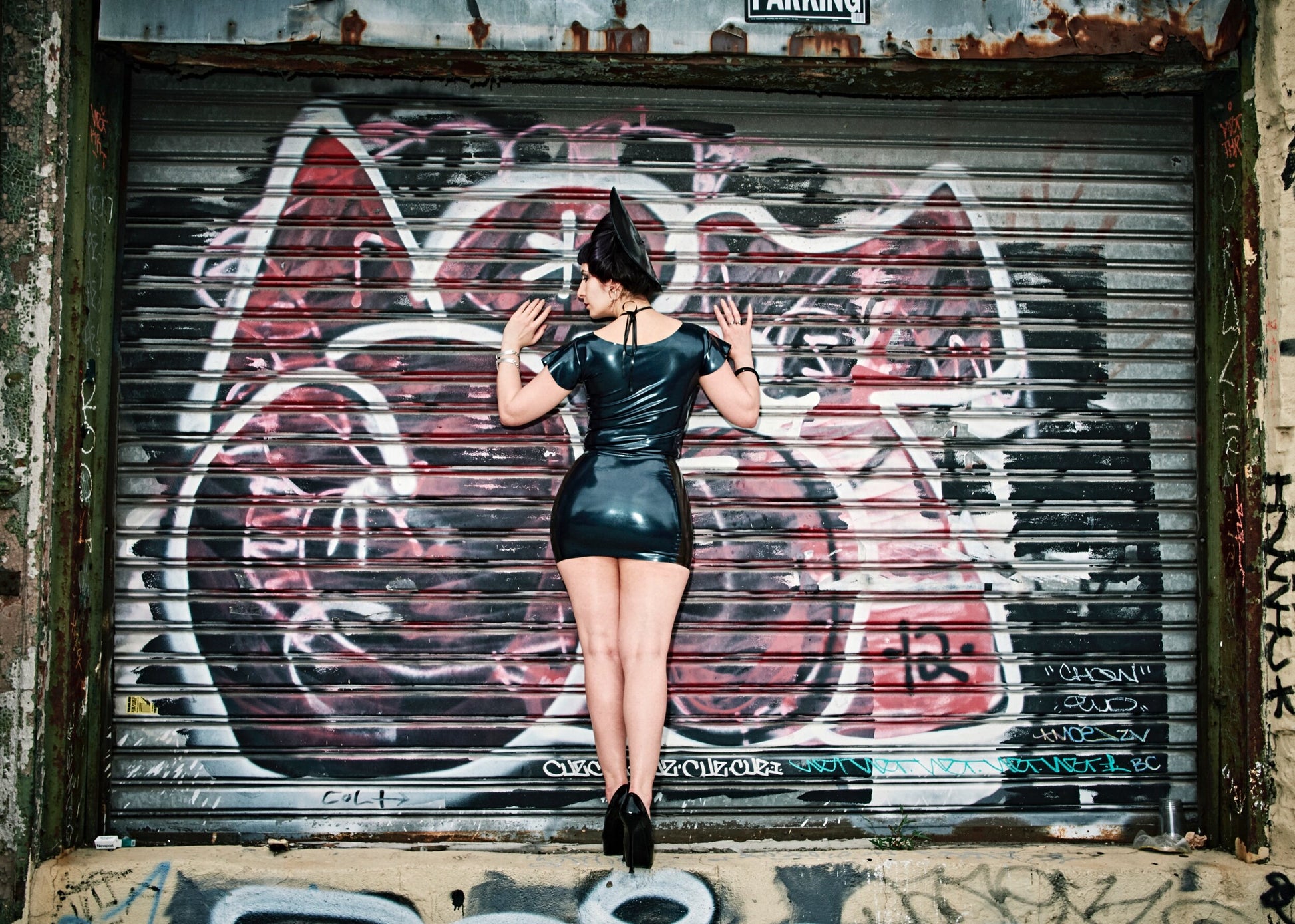 A model standing in front of a graffitied garage door showing the back of the black and black transparent Latex Girdle Mini Skirt with matching short sleeved latex top.