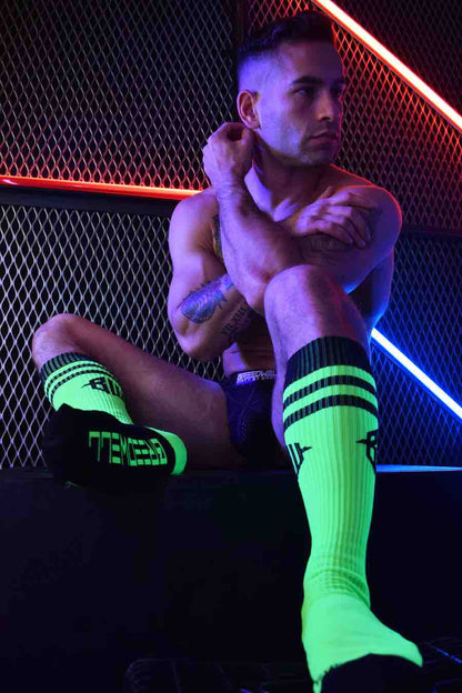 A model in a jock strap sitting in front of a fence with neon lights wearing the neon green Hex Socks.