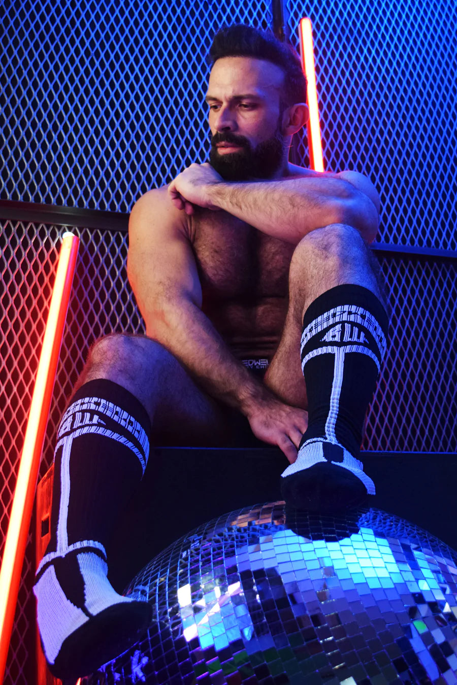 A scantily clad masculine model sits on a black box against a metal fence with neon lights. His feet sport black Hybred Socks and rest on a disco ball.
