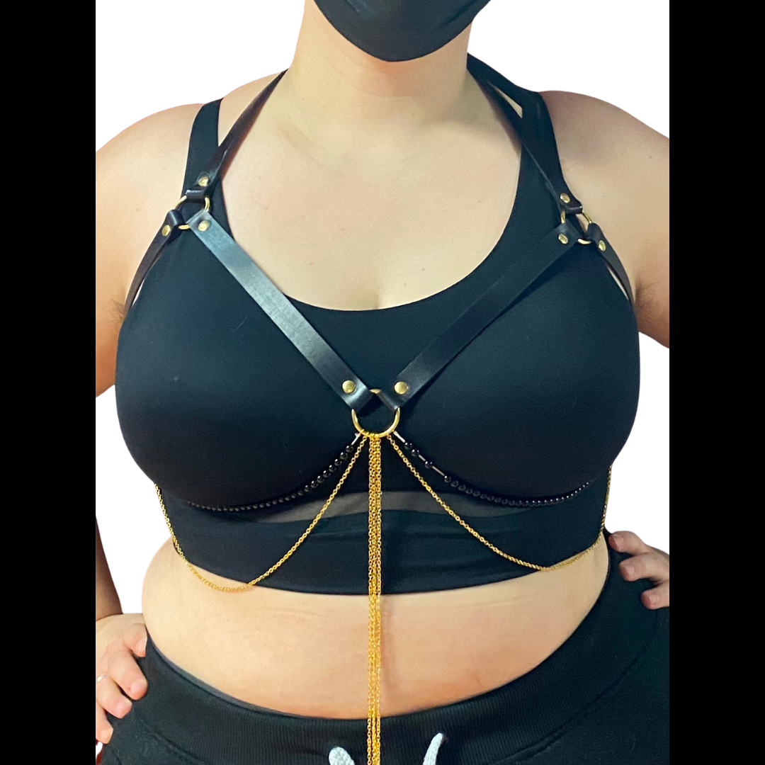 A curvy model in a black bra models the Jewel Harness in Onyx from the front.  The leather straps form a halter that frames their breasts, with two strings of black beads and several fine gold-colored chains hanging from the center ring and tracing the underside of their breasts. It fastens with gold-colored hardware.
