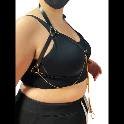 A curvy model in a black bra models the Jewel Harness in Onyx from the side. The leather straps form a halter that frames their breasts, with two strings of black beads and several fine gold-colored chains hanging from the center ring and tracing the underside of their breasts. It fastens with gold-colored hardware.