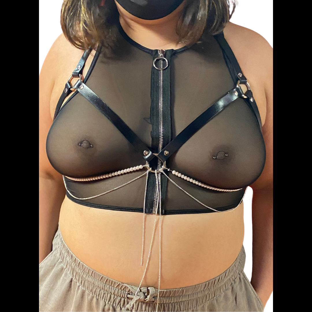 A curvy model in a transparent mesh top with pierced nipples models the Jewel Harness in Pearl from the front. The leather straps form a halter that frames their breasts, with two strings of white beads and several fine silver-colored chains hanging from the center ring and tracing the underside of their breasts. It fastens with silver-colored hardware.