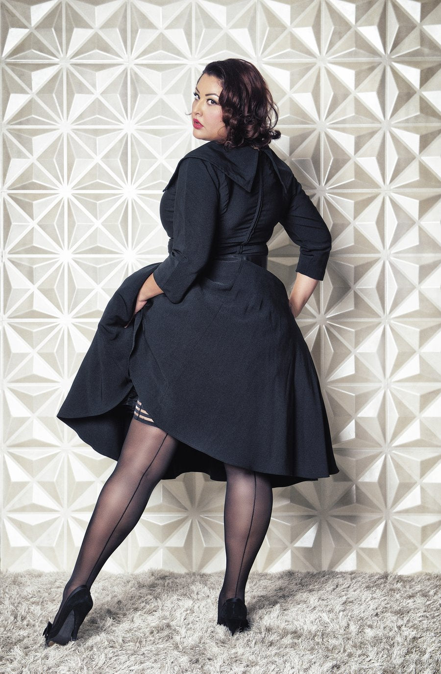 A feminine model wearing a black dress stands facing a wall, showing the back of the Lois Black Sheer Thigh Highs with Back Seam.
