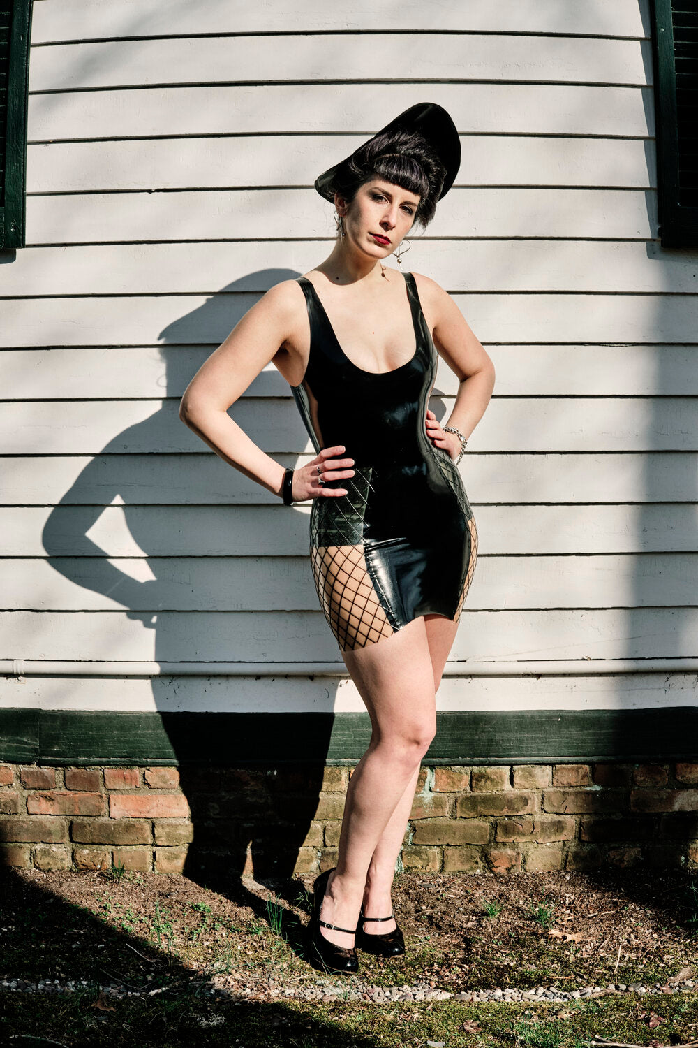 A model standing outside against a wall wearing a black latex tank top and the black and transparent fishnet Latex Specialty Girdle Skirt.
