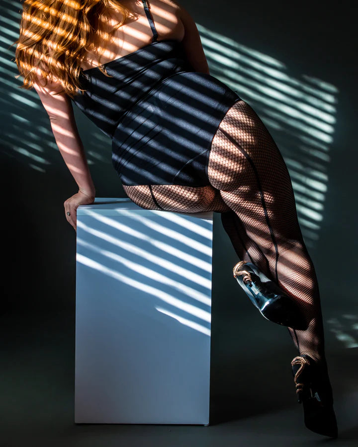 A feminine model sitting on the edge of a white table wearing a black bodysuit and the Fishnet Tights with Back Seam.