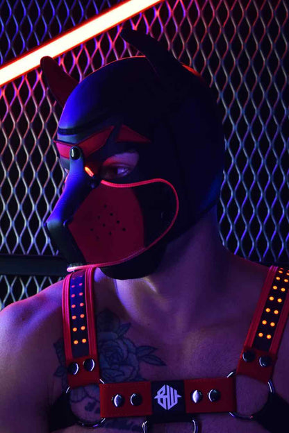 A model in a red chest harness wears the red Poundtown Glow Pup Hood in a dimly lit setting.
