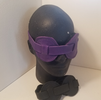 A purple round mask on a black styrofoam head. A black round blindfold lays at the base of the styrofoam head.