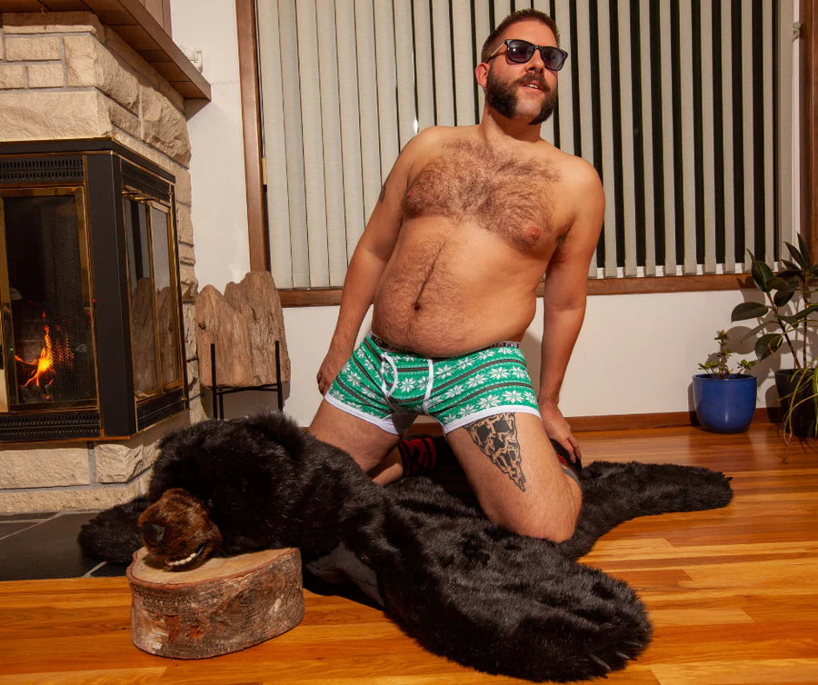 A masculine looking shirtless model kneeling on a bearskin rug wears the Winter Sweater Boxer Briefs.