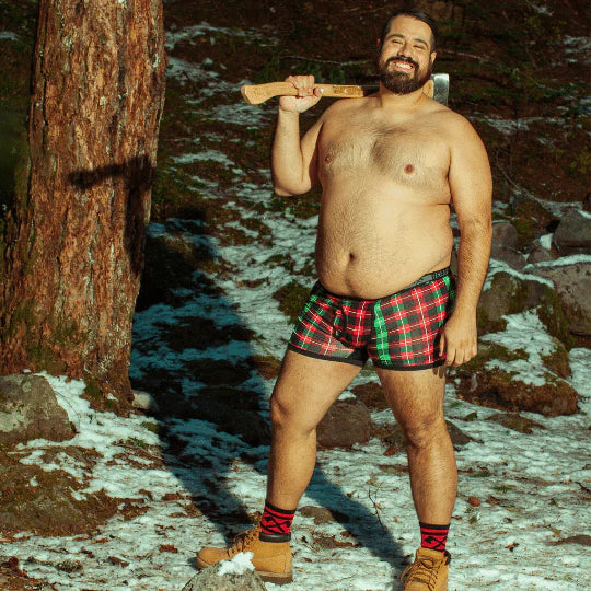 A masculine looking model stands in the snow covered forest shirtless, holding an ax and wearing the Holiday Backwoods Boxer Briefs.