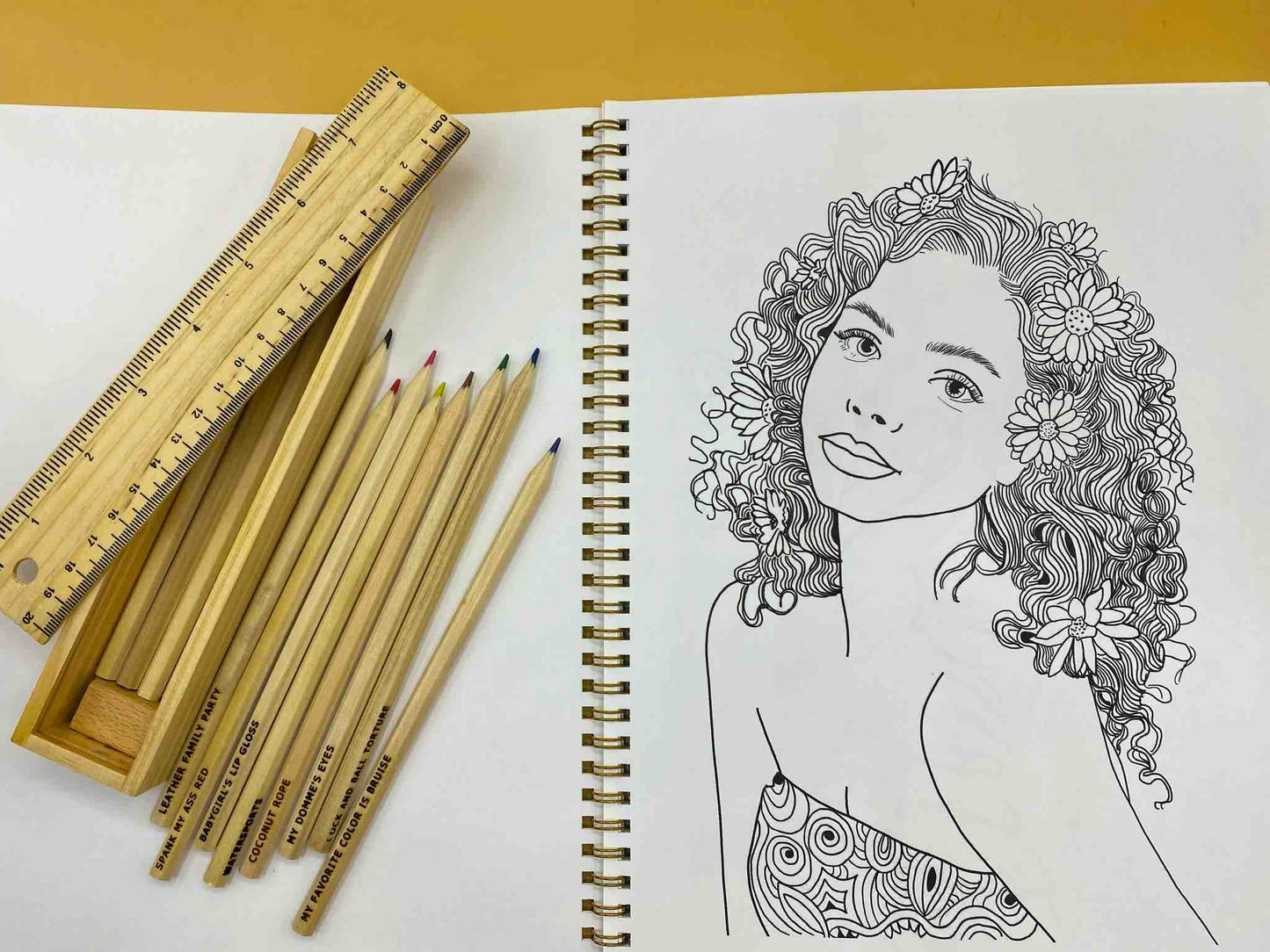 A page from Naughty and Nice: A Coloring Book for Babygirls: A girl in a tube top with flowers in her long wavy hair. Sitting atop the left page of the book is a box of colored pencils and a ruler.