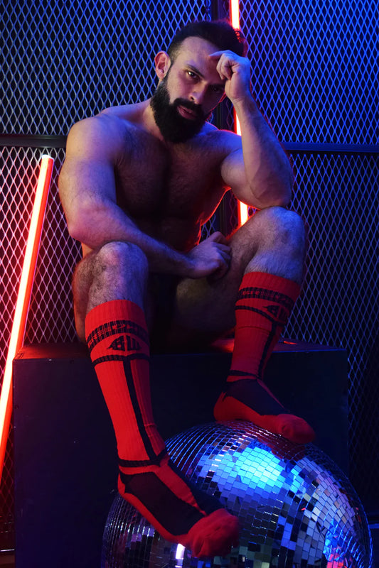 A scantily clad masculine model sits on a black box against a metal fence with neon lights. His feet sport red Hybred Socks and rest on a disco ball.